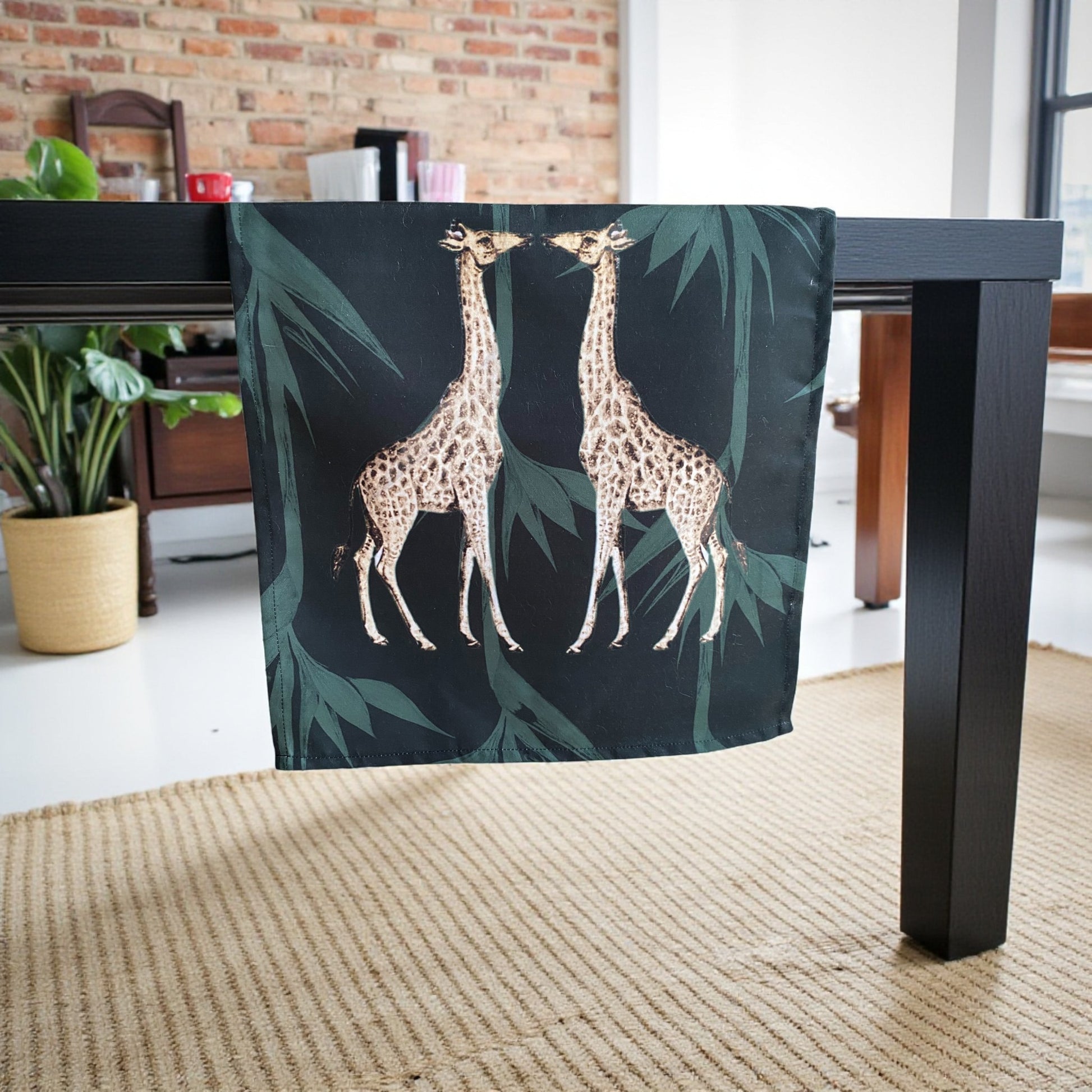 Table with a decorative tablecloth featuring giraffes and leaves in a room with plants and furniture.